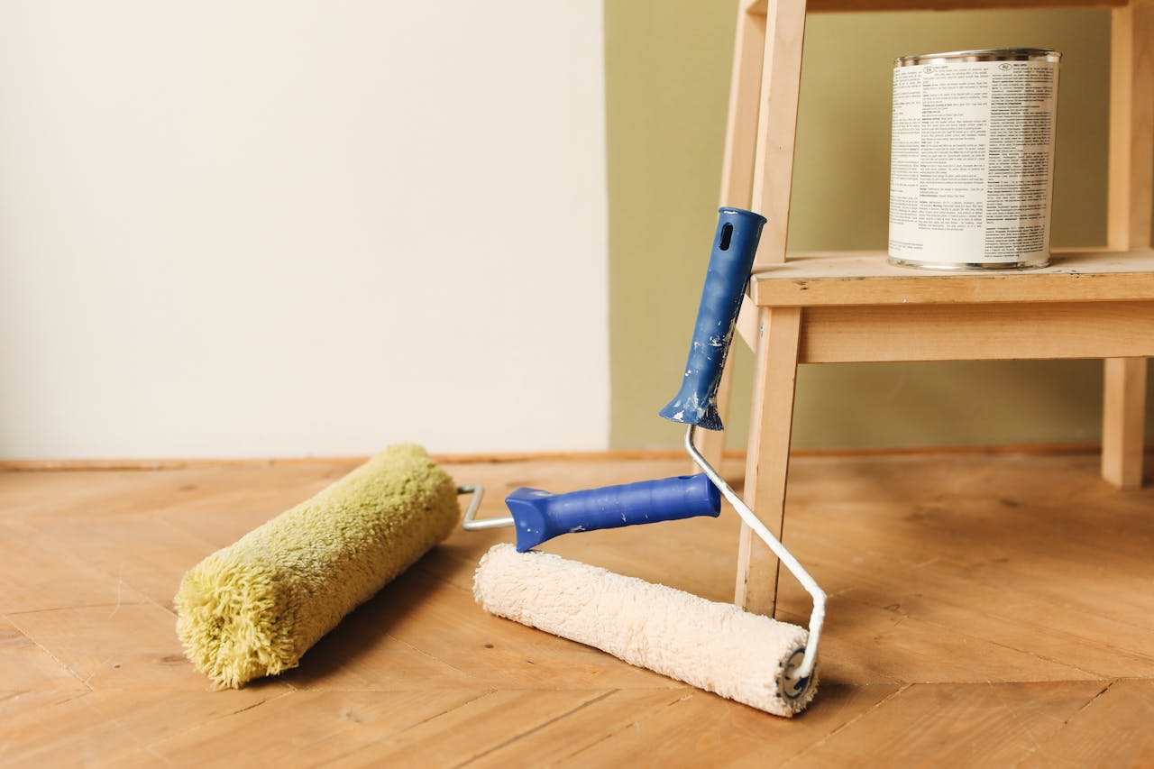 Deux rouleaux à peinture posés sur un parquet, pot de peinture sur un escabeau en bois, mur peint en deux teintes