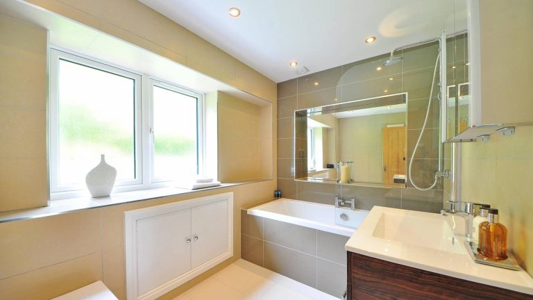 Salle de bain carrelée en grès beige, baignoire équipée d'une douche, grand miroir et meuble vasque en bois