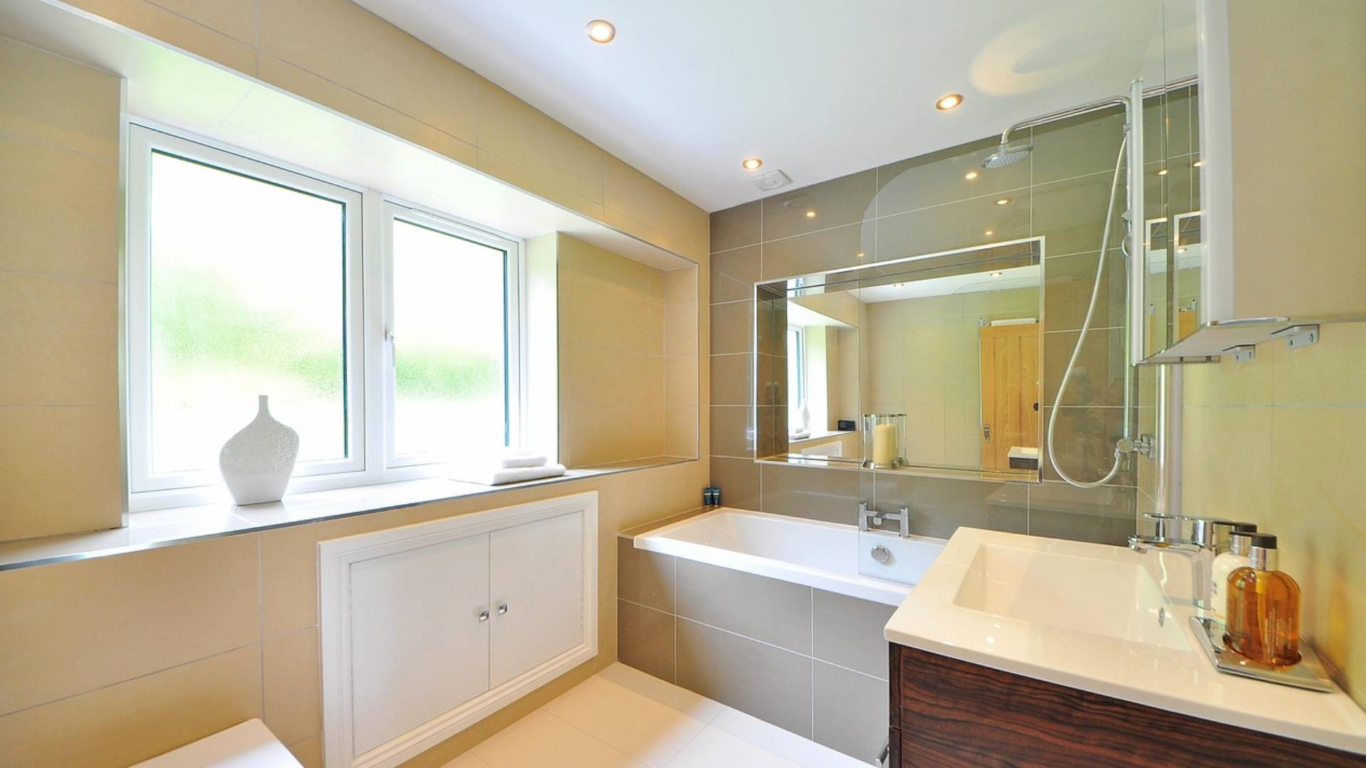 Salle de bain carrelée en grès beige, baignoire équipée d'une douche, grand miroir et meuble vasque en bois